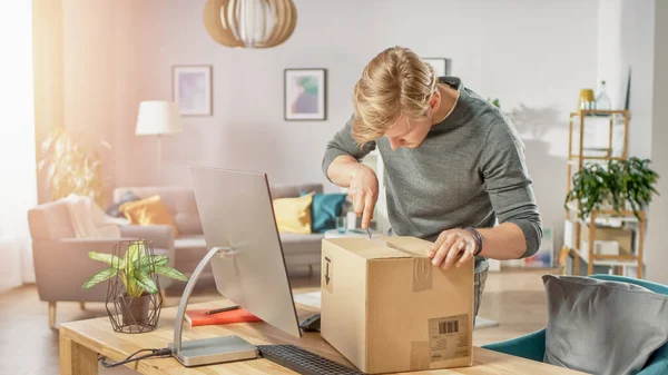 Handsome Young Man in Living Room with Cardboard Box Package, Opening it With Interest, Using Knife.