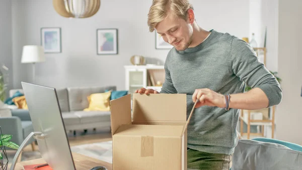 Knappe jongeman in de woonkamer opent kartonnen doos pakket met rente. — Stockfoto