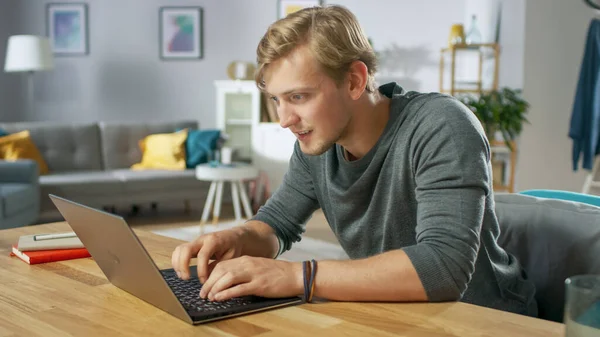 Yakışıklı genç adam rahat oturma odasının masasında otururken dizüstü bilgisayar kullanıyor.. — Stok fotoğraf