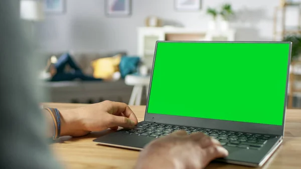 Over the Shoulder Shot of Mans Hands Using Laptop with Green Mock-up Screen. In the Background Cozy Living Room with Woman Relaxing on a Sofa. — Stock Photo, Image