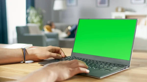 Sobre el tiro de hombro de manos humanas escribiendo en un ordenador portátil con pantalla falsa verde. En el fondo Acogedora sala de estar con mujer Relajándose en un sofá. —  Fotos de Stock