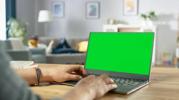 Nad ramenem záběru Mans Hands psaní na notebook s Green Mock-up Screen. V pozadí útulný obývací pokoj se ženou relaxační na pohovce. — Stock fotografie