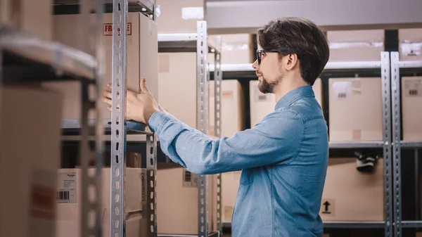 Warehouse Worker Puts Cardboard Box on a Shelf. 배경에는 상자들과 꾸러미들이 가득 들어 있고 여행 준비가 되어 있는 제품들이 가득 한 조개 집들이 있다. — 스톡 사진