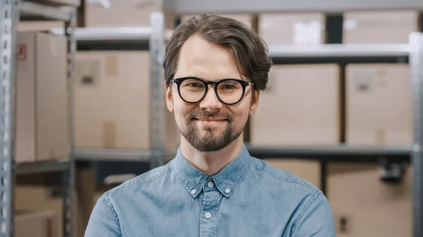 Portrait of the Handsome Warehouse Inventory Manager Standing and Smiling at the Camera. Smart Man Wearing Glasses with Rows of Shelves Full of Cardboard Boxes and Parcels Ready for Shipment. — Stock Photo, Image