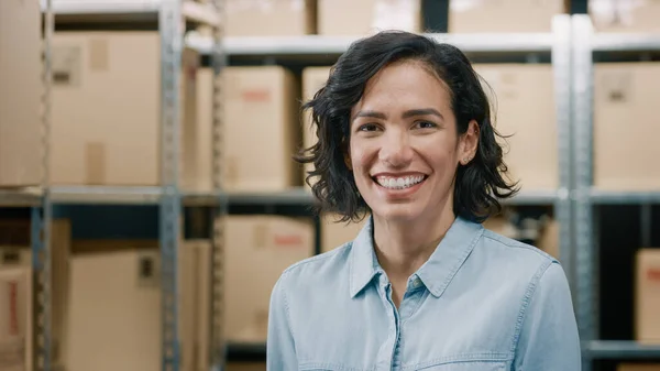 Retrato de la hermosa mujer gerente de inventario de almacén de pie y sonriendo encantadoramente. Talentosa mujer de carrera posando con filas de estantes llenos de cajas de cartón y paquetes listos para el envío. — Foto de Stock