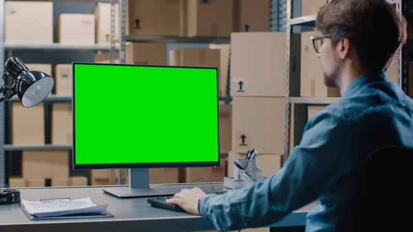 Le gestionnaire d'inventaire de l'entrepôt travaille sur un ordinateur écran vert maquillé tout en étant assis à son bureau. En arrière-plan Des étagères pleines de boîtes en carton prêtes à être expédiées. — Photo