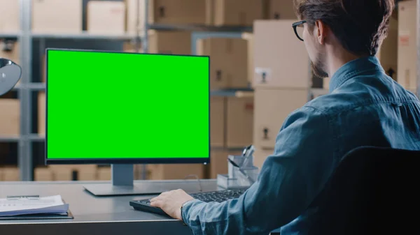 Warehouse Inventory Manager arbeitet an einem grünen Bildschirmcomputer, während er an seinem Schreibtisch sitzt. Im Hintergrund Regale voller versandfertiger Kartonverpackungen. — Stockfoto