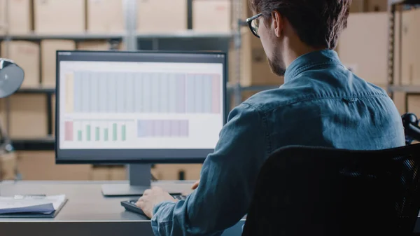 Warehouse Inventory Manager arbeitet mit einer Tabellenkalkulation auf einem Personal Computer, während er an seinem Schreibtisch sitzt. Im Hintergrund Regale voller versandfertiger Kartonverpackungen. — Stockfoto