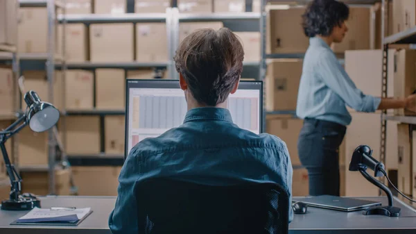 Warehouse Inventory Manager conversa com um funcionário Trabalhando com uma planilha em um computador pessoal enquanto está sentado em sua mesa. Nas prateleiras de fundo cheias de pacotes de caixa de papelão prontos para envio. — Fotografia de Stock