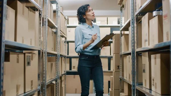 Weibliche Inventarverwalterin prüft den Bestand und schreibt in die Zwischenablage. Schöne Frau arbeitet in einem Lagerraum mit Regalreihen voller Kartons, Pakete, versandfertige Pakete. — Stockfoto