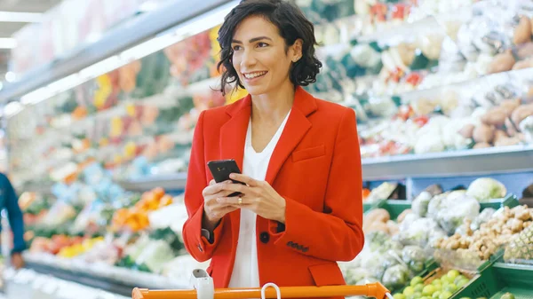 На супермаркеті: красива молода жінка використовує смартфон стоїть у секції свіжих продуктів магазину . — стокове фото