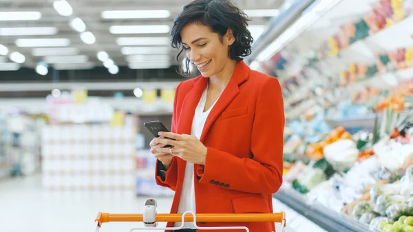 Στο supermarket: Beautiful Young Woman Χρησιμοποιεί Smartphone Ενώ Στέκεται στο τμήμα Fresh Produce του καταστήματος. — Φωτογραφία Αρχείου