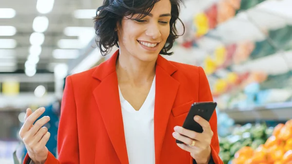 No supermercado: Retrato de uma bela jovem mulher feliz usa smartphone enquanto está na seção de produtos frescos da loja. — Fotografia de Stock