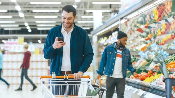 No supermercado: Jovem Bonito Usa Smartphone enquanto Empurra Carrinho de Compras e Escolhendo Legumes na Seção de Produtos Frescos. Outros clientes em segundo plano. — Fotografia de Stock