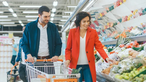 V supermarketu: Šťastný mladý pár si vybere ekologickou zeleninu v sekci čerstvé produkce obchodu. Přítel tlačí nákupní košík, zatímco přítelkyně vyzvedává potraviny. — Stock fotografie