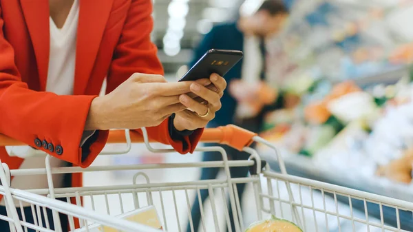 No supermercado: Mulher usa Smartphone, inclina-se no carrinho de compras. No Big Mall Woman a navegar na Internet no seu telemóvel. Concentre-se em Mãos Segurando Telefone Móvel. — Fotografia de Stock