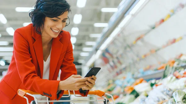 Στο Supermarket: Beautiful Smiling Woman Χρησιμοποιεί Smartphone, Leans on Shopping Cart στο Fresh Produce Section του Καταστήματος. Στο μεγάλο εμπορικό κέντρο γυναίκα περιήγηση στο Internet στο κινητό της τηλέφωνο. — Φωτογραφία Αρχείου