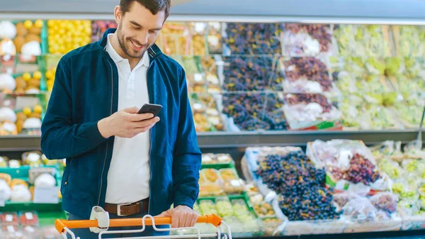 Au supermarché : Handsome Man utilise un smartphone alors qu'il se trouve dans la section des produits frais du magasin. Homme plongé dans Internet Surfer sur son téléphone portable En arrière-plan Fruits colorés et — Photo