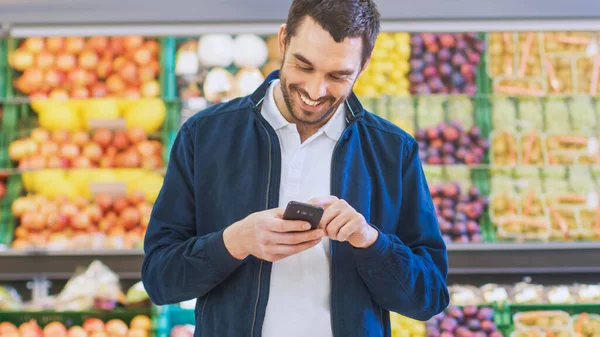Στο σούπερ μάρκετ: Όμορφος άνθρωπος χρησιμοποιεί Smartphone, ενώ στέκεται στο Fresh Produce Τμήμα του καταστήματος. Άνθρωπος βυθισμένο στο Internet Surfing στο κινητό του τηλέφωνο στο παρασκήνιο πολύχρωμα φρούτα και — Φωτογραφία Αρχείου