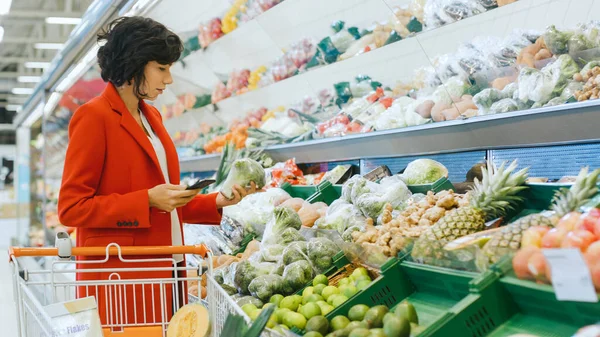 No supermercado: Tiro de uma bela jovem mulher caminha através de produtos frescos seção, escolhe brócolis e coloca-los em seu carrinho de compras. — Fotografia de Stock
