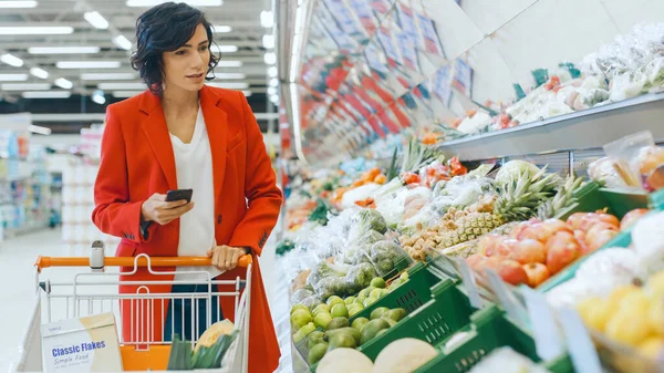 Στο supermarket: Beautiful Young Woman Walks Through Fresh Produce Section, Επιλέγει Λαχανικά και Ώθηση Καλάθι Αγορών. Πελάτης Χρησιμοποιεί Smartphone ενώ Ψώνια για Φρούτα και Λαχανικά στο — Φωτογραφία Αρχείου