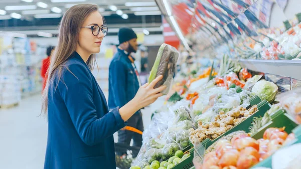 У супермаркеті: Красива молода жінка стоїть у секції свіжих продуктів, вибирає органічні овочі та фрукти. Покупки клієнтів на задньому плані . — стокове фото