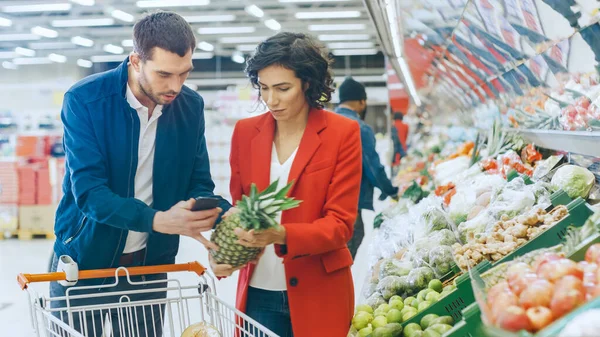 У супермаркеті: Щаслива пара робить покупки, вибираючи фрукти та овочі в секції свіжих продуктів. Чоловік використовує смартфон і натискає кошик для покупок, жінка розміщує продукти у візок . — стокове фото