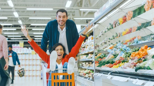 Στο Supermarket: Man Pushes Shopping Cart with Woman Sitting in it, Happy Couple Has Fun Racing in a Trolley μέσω του Fresh Produce Section του Καταστήματος. Οι άνθρωποι περπατούν. — Φωτογραφία Αρχείου