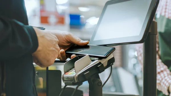 체크아웃 카운터에서 스마트 폰으로 돈을 지불하고 있는 남성의 슈퍼 마켓 근접 보도입니다. 빅 몰에서 현대적 이고 편리 한 무선 NFC 지불 시스템을 사용하는 일. — 스톡 사진