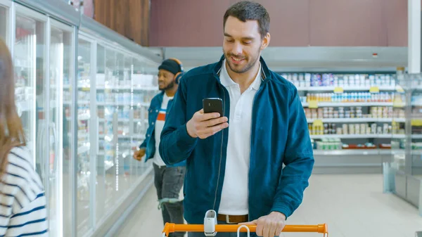 Στο σούπερ μάρκετ: Όμορφος άνθρωπος με καλάθι αγορών Χρησιμοποιεί Smartphone, Smiles and Walks Through Frozen Goods Section. Άλλοι πελάτες Αγορές στο παρασκήνιο. — Φωτογραφία Αρχείου