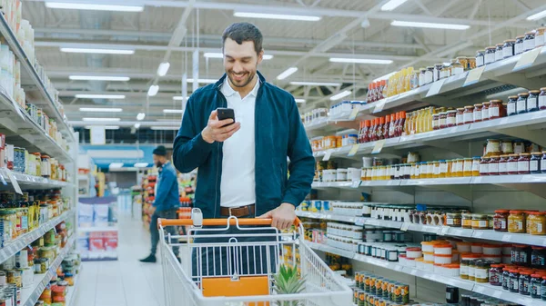 I snabbköpet: Stilig Man Använder Smartphone, Le när du står på avdelningen Konserverade varor. Har kundvagn med hälsosamma matvaror inuti. Andra kunder Promenader i bakgrunden. — Stockfoto