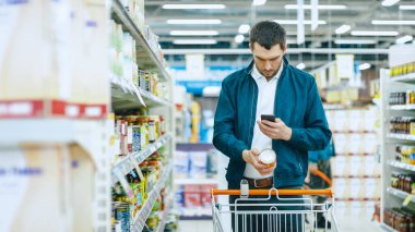 Süpermarkette: Yakışıklı Adam Akıllı Telefon Kullanıyor ve Malzeme Kutusunun Resmini Çekiyor. Konserve Ürünler Bölümünde Alışveriş Arabasıyla Duruyor.