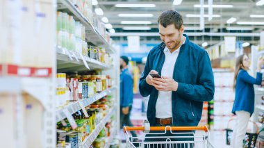 Süpermarkette Yakışıklı Adam Akıllı Telefon Kullanıyor ve Konserve Gıda Bölümü 'nde Stands. En Sağlıklı Ürün Arıyor.