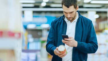 Süpermarkette: Yakışıklı Adam Akıllı Telefonu Konserve Mallarının Beslenme Değerlerini Kontrol Etmek İçin Kullanıyor ve Satın Alıyor. Konserve Ürünler Bölümünde Alışveriş Arabasıyla Duruyor.