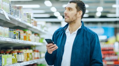 Süpermarkette Yakışıklı Adam Akıllı Telefon Kullanıyor, Konserve Ürünler Bölümü 'nde Dururken Gülümsüyor.