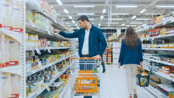 Im Supermarkt: Schöner Mann durchstöbert Regal mit Konserven, schaut auf Blechdose, entscheidet sich aber gegen den Kauf. Er läuft mit dem Einkaufswagen durch verschiedene Bereiche des Ladens. — Stockfoto