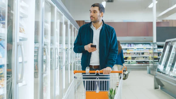 Im Supermarkt: Schöner Mann benutzt Smartphone, lächelt und geht durch Tiefkühlkost Andere Kunden kaufen im Hintergrund ein. — Stockfoto