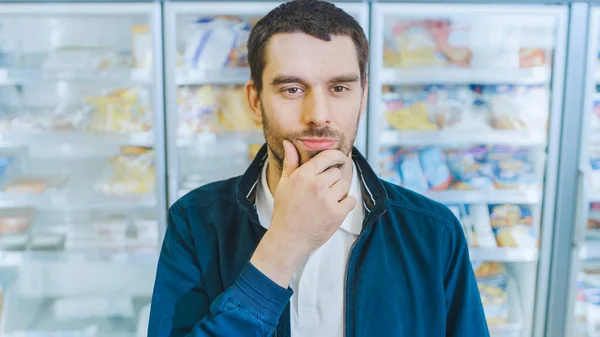 Portrait Shot of the Handsome Man Thiking about Choosing Tin Can from the Canned Goods Section. In the Background Frozen Goods Section of the Store.