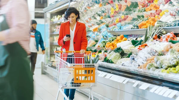 No supermercado: Mulher bonita com carrinho de compras usa smartphone e caminha através de produtos frescos seção da loja. — Fotografia de Stock