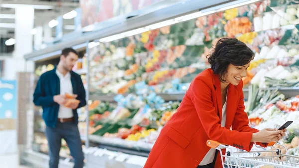 No supermercado: bela jovem mulher usa smartphone enquanto está de pé na seção de produtos frescos da loja. No fundo Outros Clientes Compras. — Fotografia de Stock