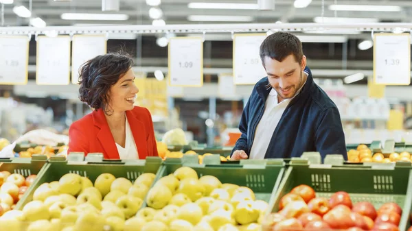 En el supermercado: Feliz pareja joven elige fruta orgánica en la sección de productos frescos de la tienda. Esposo utiliza Smartphone, Esposa recoge fruta. — Foto de Stock