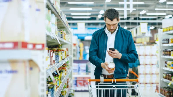 I snabbköpet: Stilig man använder smartphone och tar bild av burken av varor. Hes Stående med kundvagn i Konserverade varor sektionen. — Stockfoto