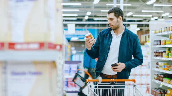 Im Supermarkt: Schöner Mann nutzt Smartphone und stöbert im Konservenregal Hes steht mit Einkaufswagen in Konservenabteilung. — Stockfoto