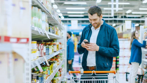 Στο σούπερ μάρκετ: Όμορφος άνθρωπος χρησιμοποιεί Smartphone και στέκεται στο τμήμα κονσερβοποιημένων προϊόντων. Ψάχνει για ένα καλύτερο υγιεινό προϊόν. — Φωτογραφία Αρχείου