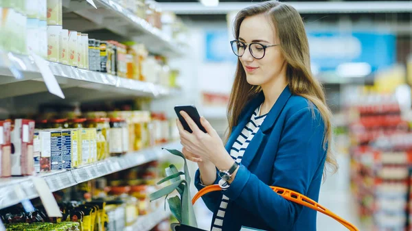 No supermercado: bela jovem mulher usa smartphone enquanto navega pela seção de produtos enlatados da loja. Ela verifica sua lista de compras e mantém a cesta de compras. — Fotografia de Stock