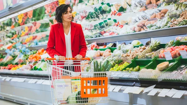 У супермаркеті: красива молода жінка проходить через секцію свіжих продуктів, вибирає овочі та кошик для покупок. покупки фруктів і овочів в магазині . — стокове фото
