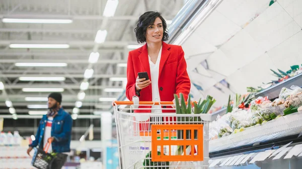 No supermercado: Mulher bonita com carrinho de compras usa smartphone e caminha através de produtos frescos seção da loja. — Fotografia de Stock