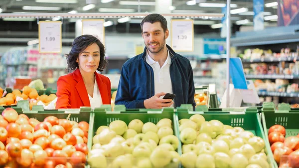 У супермаркеті: Щаслива молода пара вибирає органічні фрукти в секції свіжих продуктів магазину. Чоловік використовує смартфон, дружина підбирає фрукти . — стокове фото
