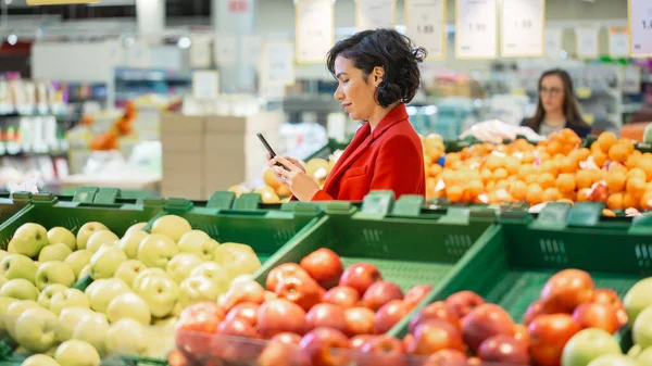 No supermercado: Mulher bonita usa smartphone enquanto está na seção de produtos frescos da loja. Mulher imersa na Internet Surfando em seu telefone celular no fundo Frutas coloridas — Fotografia de Stock