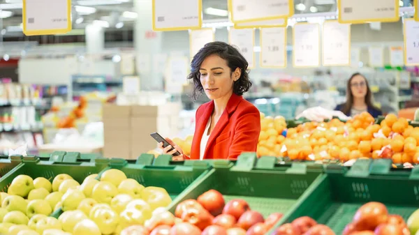 No Supermercado: Mulher Bonita Escolhe Frutas Orgânicas na Seção de Produtos Frescos do Mercado de Agricultores. Ela pega frutas e segura Smartphone em outra mão. — Fotografia de Stock